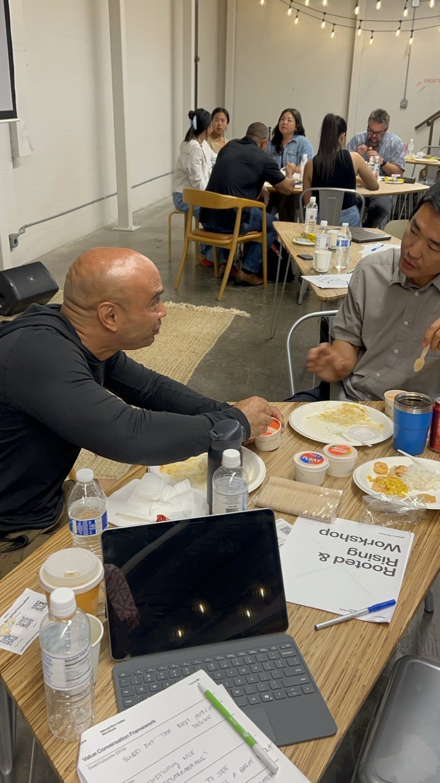 Two men talking at a table with laptops and a workbook